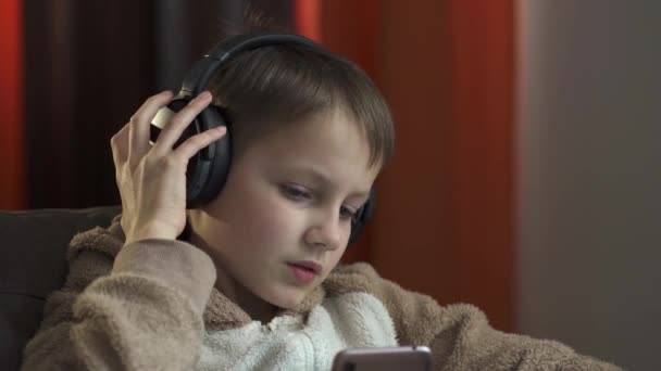 Facet w słuchawkach i ze smartfonem w rękach siedzi na kanapie i słucha muzyki. Kwarantanna 2020 — Wideo stockowe