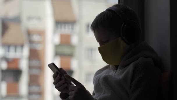 El niño con una máscara protectora se sienta en un alféizar de la ventana con el teléfono en las manos. Karatnin. — Vídeo de stock