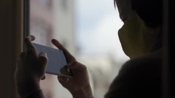 Teenager v ochranné masce hrát hry na smartphone zatímco sedí na parapetu. Karatnin 2020 — Stock video