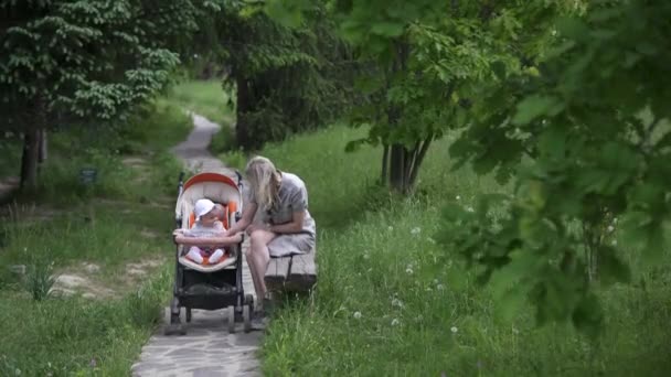 Mamma med en liten dotter som vilar i en park på en bänk. Ett år gammalt barn sitter i en vagn och leker. — Stockvideo