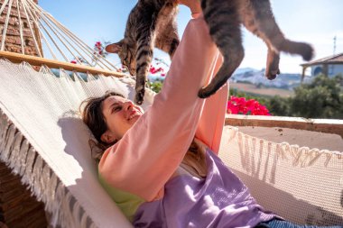 Genç İspanyol kadın beyaz hamakta tekir kedisiyle birlikte evdeki terasta yatıyor.