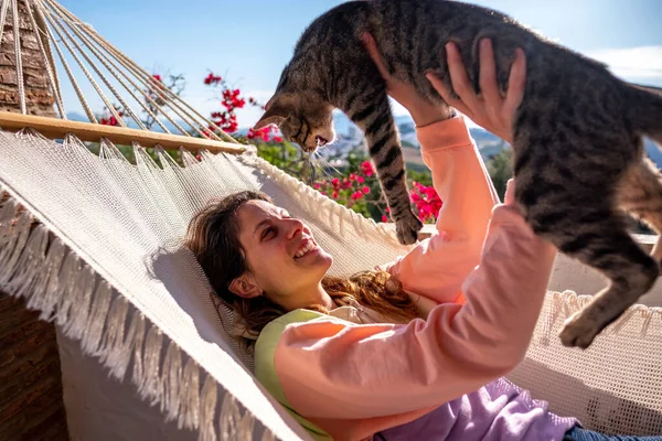 Genç İspanyol kadın beyaz hamakta tekir kedisiyle birlikte evdeki terasta yatıyor.