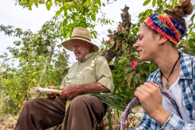 Hasır şapkalı yaşlı adam kütüğün üzerinde sebze keserken torunu ona yardım ediyor.