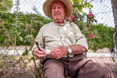 Hasır şapkalı yaşlı adam kütüğün üzerinde oturmuş sebze kesiyor.