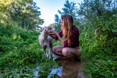 Genç bir kadın uzun yeşil otlarla çevrili köpeğiyle nehirde yürüyüş yapıyor.