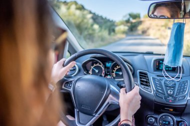 Dikiz aynasından sarkan yüz maskeli genç Latin kadın arabasını kullanıyor.