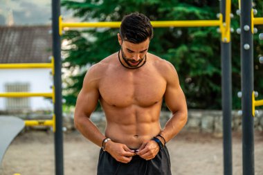 Bir spor salonunun barında, gömleksiz, siyah pantolonsuz ve maske takan genç bir Latin adamın yakın portresi.