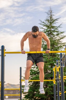 Tişörtsüz, siyah pantolonlu ve maskeli genç bir adam spor salonu barında barfiks çekiyor.