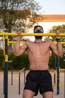 Siyah pantolonlu ve maskeli genç Latin adam jimnastik parkında tişörtsüz egzersiz yapıyor.