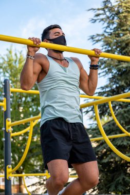 Soluk mavi tişörtlü, siyah pantolonlu ve maskeli genç bir Latin jimnastik parkında egzersiz yapıyor.