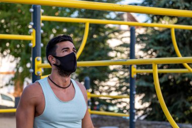 Soluk mavi tişörtlü, siyah pantolonlu ve maskeli genç bir Latin jimnastik parkında egzersiz yapıyor.