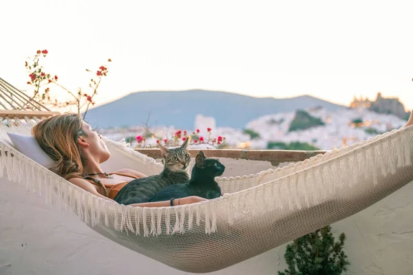 Beyaz bir hamakta iki kedisiyle dinlenen turuncu tulumlu genç Latin kadın.