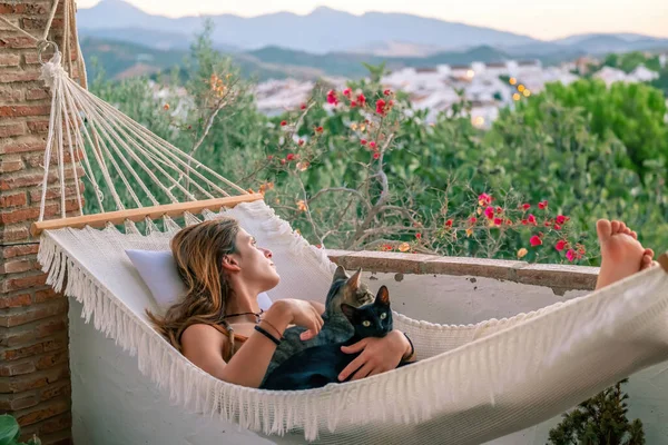 Young latin woman with orange dungaree resting in a white hammock with her two cats