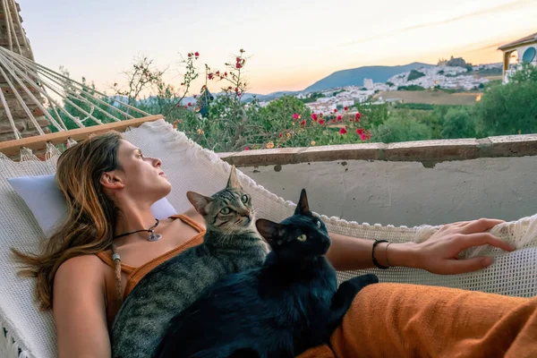 Beyaz bir hamakta iki kedisiyle dinlenen turuncu tulumlu genç Latin kadın.