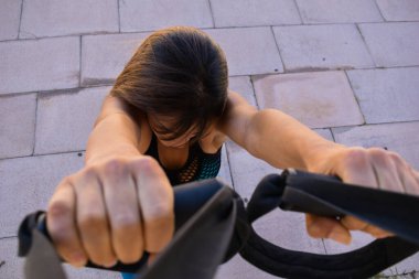 Açık havada Trx ile spor yapan ve kameraya bakan bir kadın.