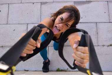 Açık havada Trx ile spor yapan ve kameraya bakan bir kadın.