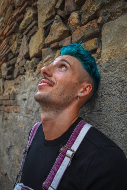 Mavi saçlı, eski tuğla bir duvara yaslanmış genç bir adamın yüzüne yakın çekim.