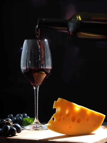 Red wine poured into a large wine glass from a dark bottle on a black background with black grapes and cheese