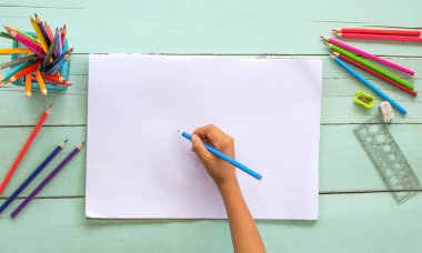 Tahta bir masanın üzerine beyaz kağıt üzerine çizilmiş renkli kalemler kullanan bir çocuğun elinin en üst görüntüsü..