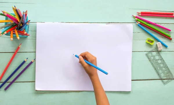 Tahta bir masanın üzerine beyaz kağıt üzerine çizilmiş renkli kalemler kullanan bir çocuğun elinin en üst görüntüsü..
