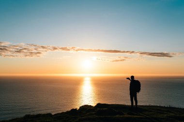 Sahilde gün batımını seyreden ve telefon kamerasıyla fotoğraf çeken genç bir yürüyüşçünün silueti. Seyahat kavramı. 