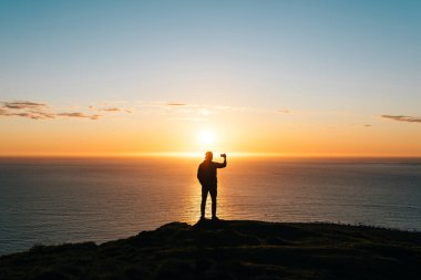 Deniz kenarında gün batımını izleyen ve telefon kamerasıyla fotoğraf çeken genç bir adamın silueti. Seyahat kavramı. 