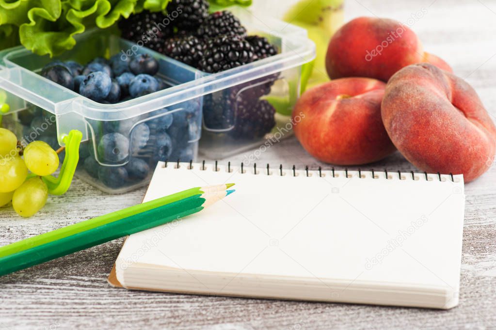 Empty notebook and berries in lunch box and fruits and vegetables on wooden table