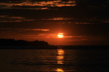 Koyu kırmızı gün batımı bulutların arkasından batıyor ve su yüzeyindeki ışık yansıması, tropikal plajda, Gili Hava Adası, Lombok, Endonezya