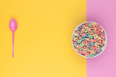 Bir kase renkli mısır gevreği ve kaşık üst görünümü. Metin için boş yer var. Metin için boş yer var. İki renkli arkaplan, sarı ve pembe.