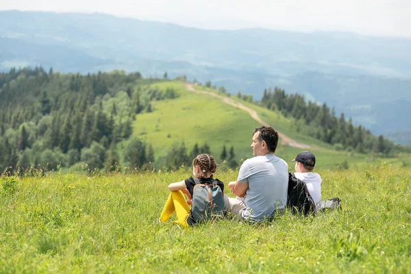 Baba ve çocuklar dağın tepesinde oturur ve güzel bir manzaraya bakarlar. Aile gezisi