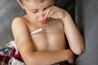 Çocuk sıcaklığı cam termometreyle ölçüyor. Arka planda bir çocuk sulu gözleri ovar. Soğuk algınlığı belirtileri.