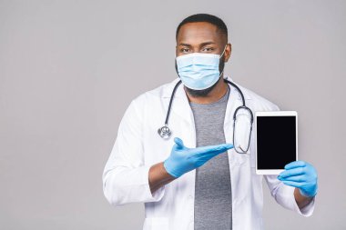 Gri arka planda izole edilmiş cerrahi hijyen koruyucu maske takan Afrikalı Amerikalı doktor ve elinde boş fotokopi ekranı tablet bilgisayarı var.. 
