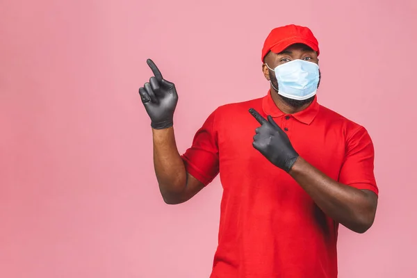 Delivery man in red cap blank t-shirt uniform sterile face mask gloves isolated on pink background. Guy employee working courier Service quarantine pandemic coronavirus virus. Pointing finger.