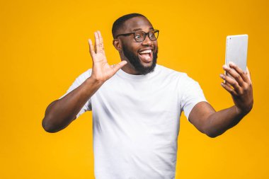 Tatildeki neşeli Afro-Amerikalı adamın portresi, sarı arka planda izole edilmiş, günlük giyinmiş, tabletinde veb kamera kullanıyor, eğleniyor, gülümsüyor..