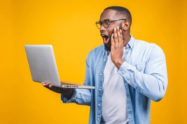 Genç sürpriz Afrikalı adam ayakta ve dizüstü bilgisayar kullanıyor sarı arkaplan üzerinde izole.