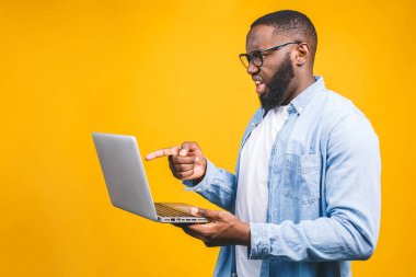 Genç sürpriz Afrikalı adam ayakta ve dizüstü bilgisayar kullanıyor sarı arkaplan üzerinde izole.