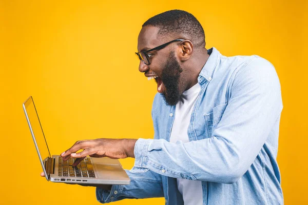 Young surprised african american man standing and using laptop computer isolated over yellow background.