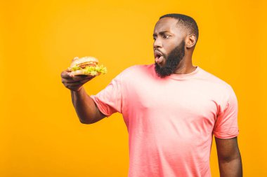 Sarı arka planda izole edilmiş hamburger yiyen Afrikalı genç bir Amerikalı..