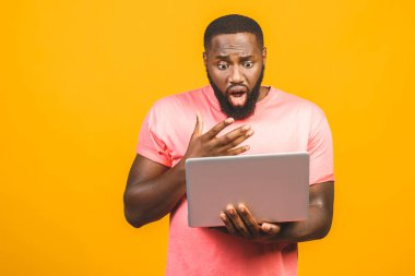 Genç sürpriz Afrikalı adam ayakta ve dizüstü bilgisayar kullanıyor sarı arkaplan üzerinde izole.