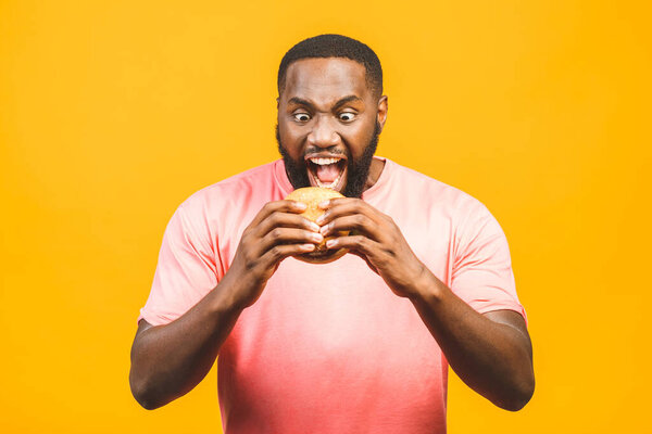 Young african american man eating hamburger isolated over yellow background.