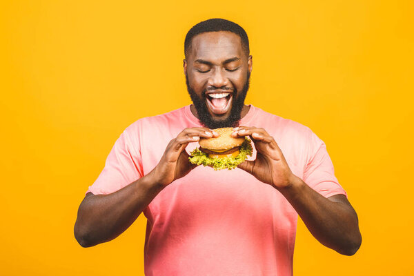 Young african american man eating hamburger isolated over yellow background.