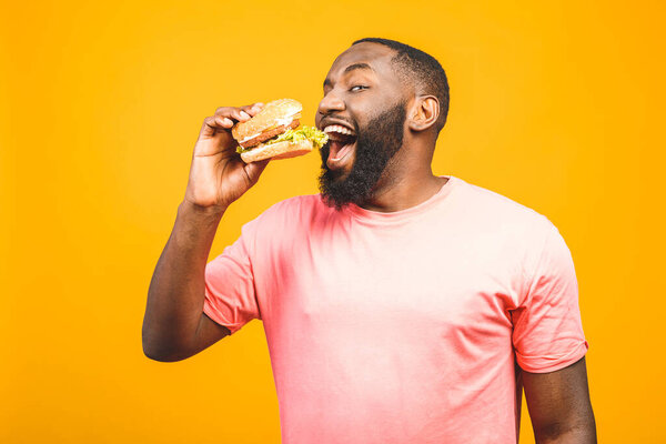 Young african american man eating hamburger isolated over yellow background.