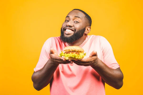 Young african american man eating hamburger isolated over yellow background.