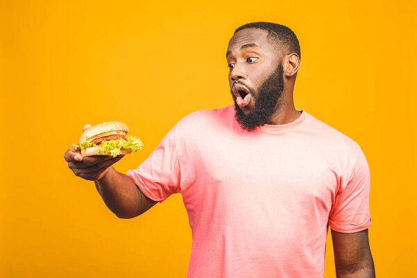 Young african american man eating hamburger isolated over yellow background.