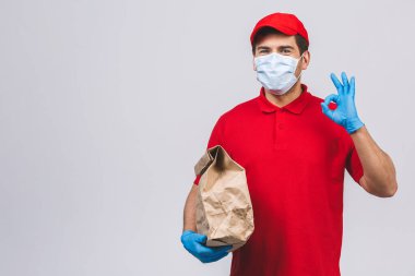 Kırmızı şapkalı, boş tişörtlü, maske eldivenli teslimatçı eleman beyaz arka planda boş karton kutu tutuyor. Servis karantina salgını Coronavirus virüsü 2019-ncov konsepti.