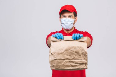 Kırmızı şapkalı, boş tişörtlü, maske eldivenli teslimatçı eleman beyaz arka planda boş karton kutu tutuyor. Servis karantina salgını Coronavirus virüsü 2019-ncov konsepti.