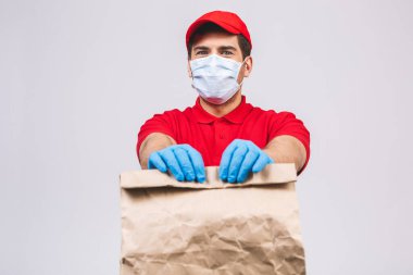 Kırmızı şapkalı, boş tişörtlü, maske eldivenli teslimatçı eleman beyaz arka planda boş karton kutu tutuyor. Servis karantina salgını Coronavirus virüsü 2019-ncov konsepti.