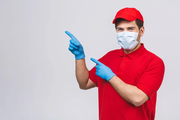 Image of optimistic delivery man in red t-shirt, mask and cap smiling and pointing finger up on copyspace text or product isolated over white background.