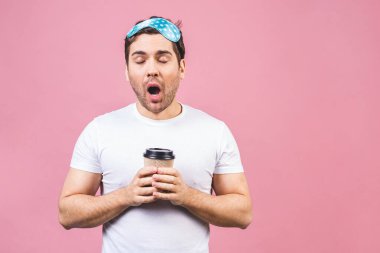 Mavi uyku maskesi ve bir fincan kahve ya da çay ile huzurlu, uykulu genç bir adamın portresi. Pembe arka planda izole edilmiş.