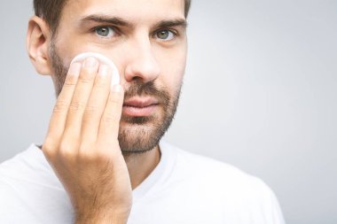 Gri arka planda pamuk pedlerle cildini temizleyen yakışıklı bir adam ve kameraya bakıyor. Yakın plan